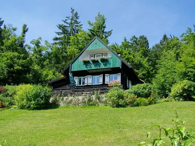 A log cabin with green metal roof surrounded by dense forest and green meadow on sunny day