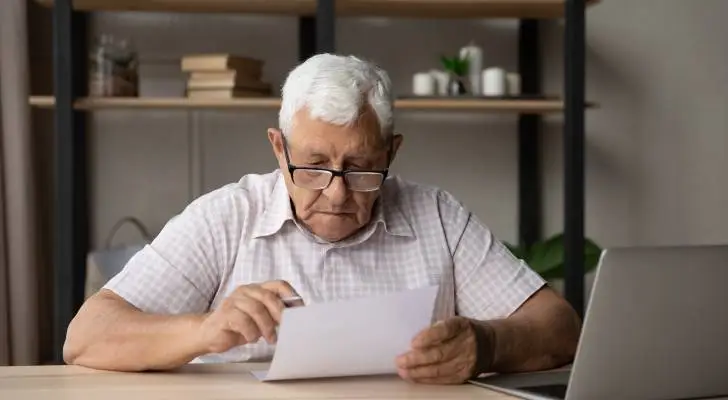 Older man with white hair and glasses sitting at desk, reviewing documents with laptop nearby