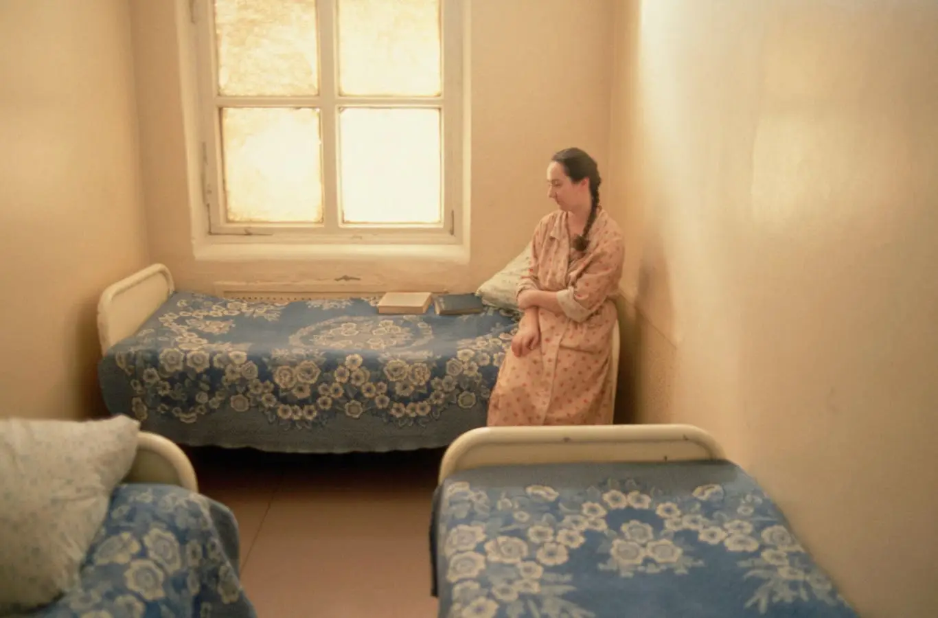 Woman in pink robe stands beside two beds with patterned blue coverlets in sparse room with window