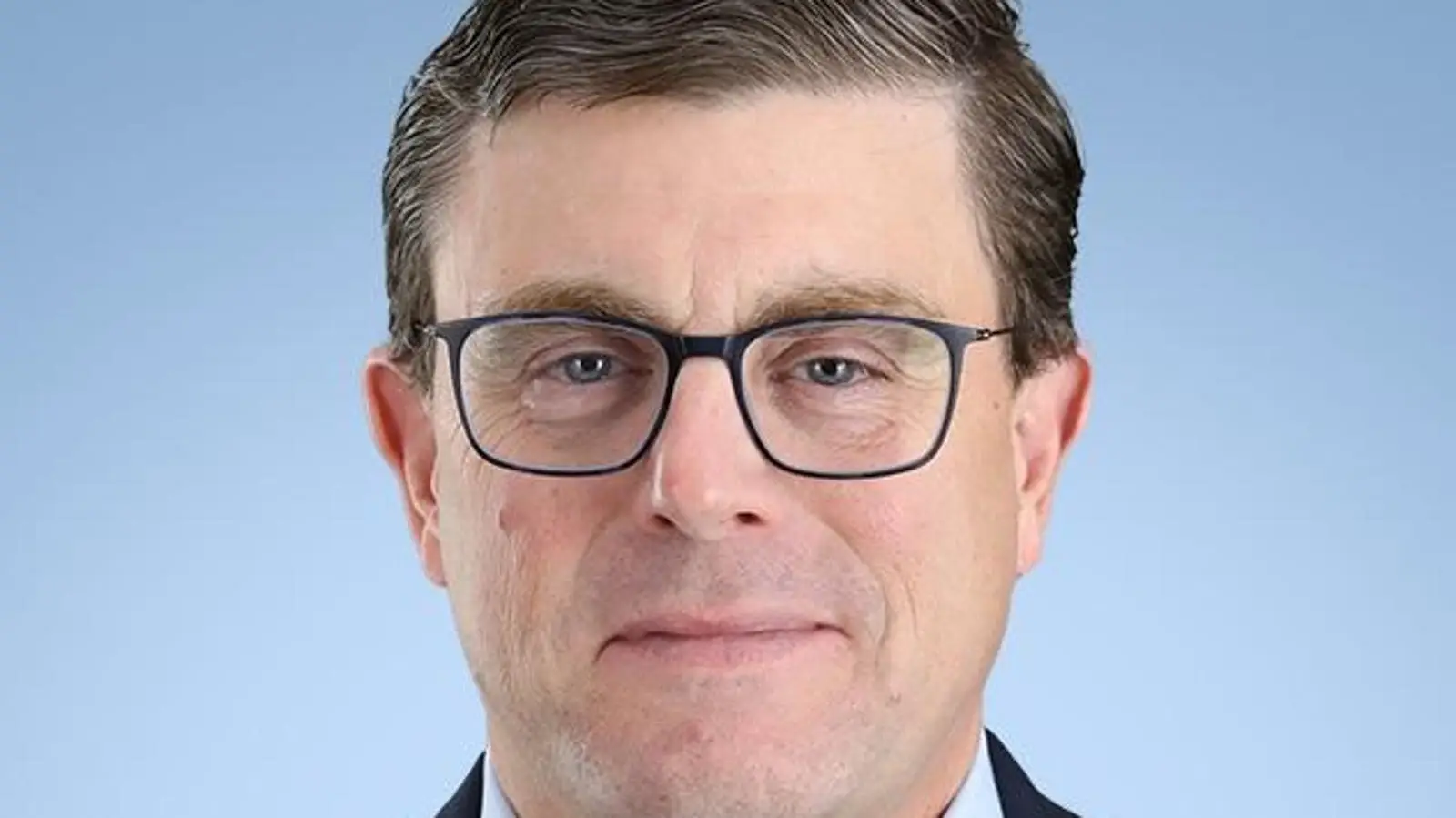 Professional headshot of a man wearing black-framed glasses and dark blazer against light blue background