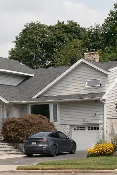 Single-story house with white siding, gray roof, attached garage, dark sedan in driveway, yellow flowers and brown shrub landscaping