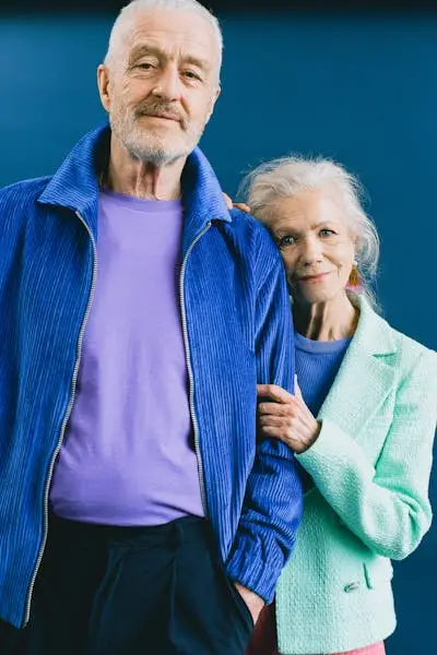 Older man in blue jacket and purple shirt stands beside older woman in mint green cardigan against blue background
