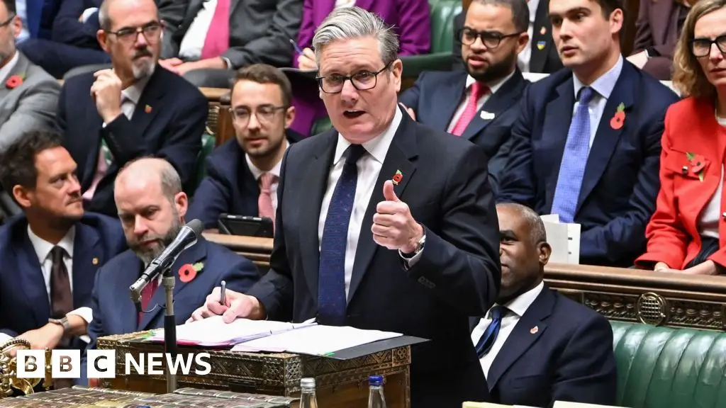 Man in dark suit and glasses speaks at parliamentary podium while colleagues stand nearby