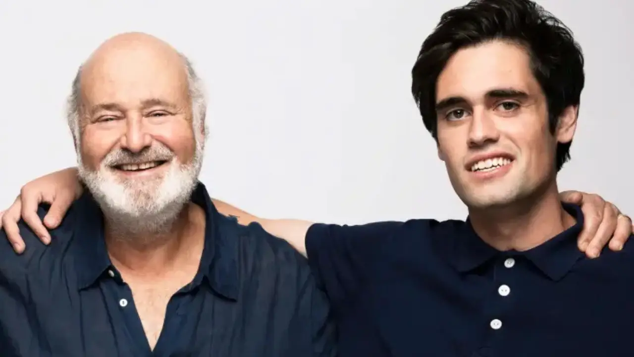 Two men in dark polo shirts stand together against a light gray background, one older with white beard, one younger with dark hair