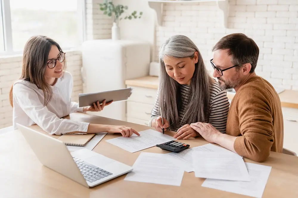 Three people review documents together at a wooden table with a laptop and calculator
