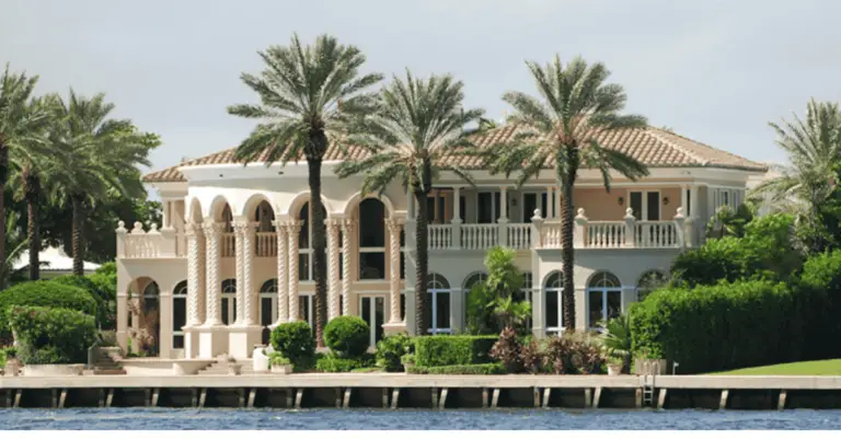Waterfront mansion with cream-colored facade, arched windows, tile roof, palm trees, and manicured hedges