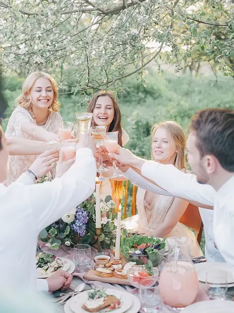 Group of five people toasting with champagne glasses outdoors under flowering tree branches at a garden table