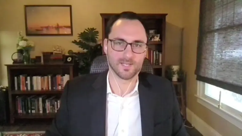 Man in glasses and dark blazer smiling at camera in home office with bookshelves and plants behind him