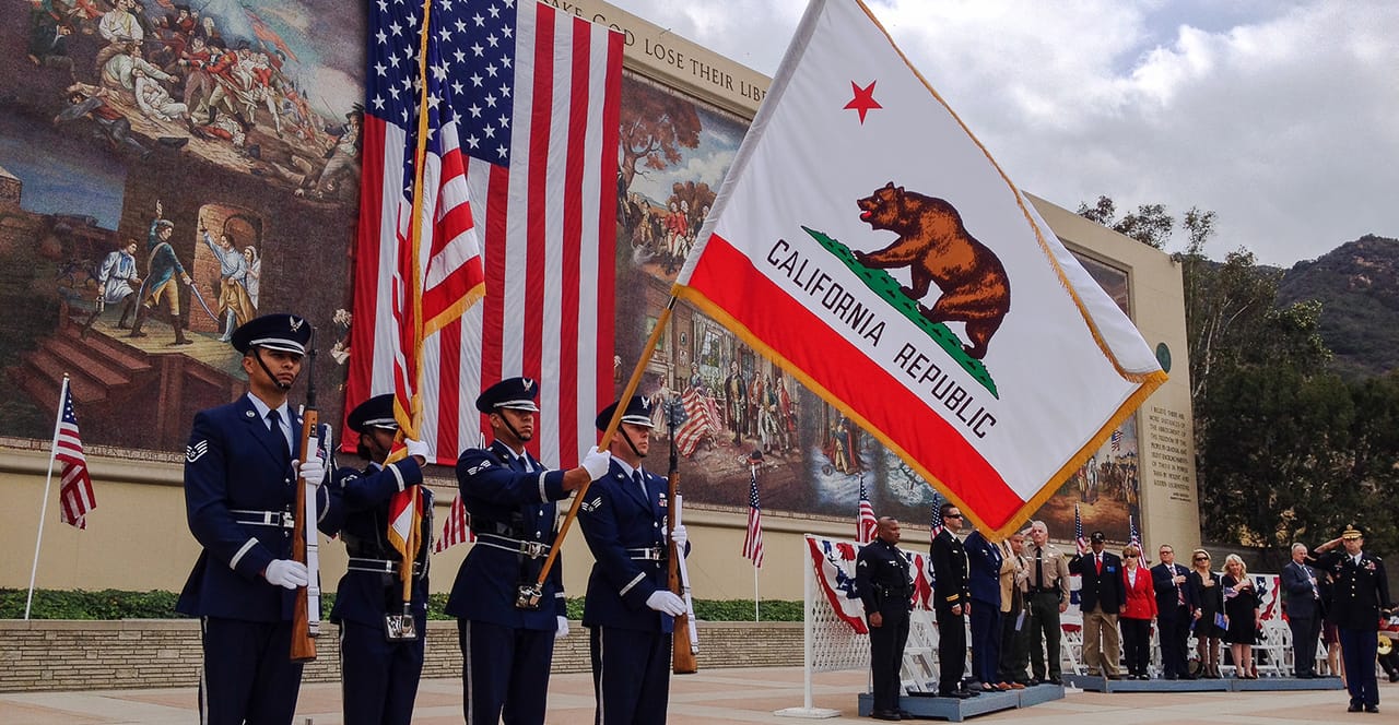 Military honor guard in dress uniforms holds American and California state flags during outdoor ceremony