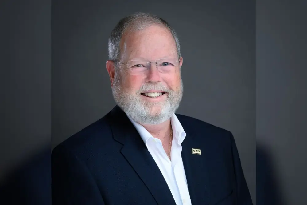 Professional headshot of an older man with gray hair and beard wearing glasses and a dark blazer over a white shirt