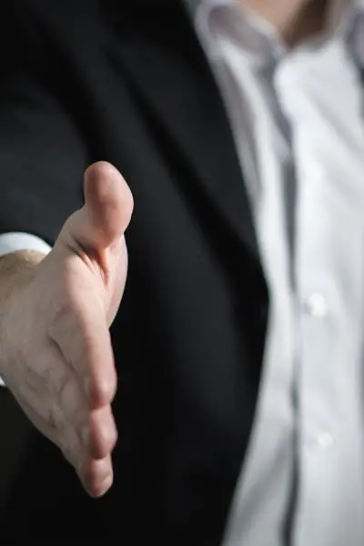 Businessman in dark suit and white shirt extends hand toward camera in handshake gesture