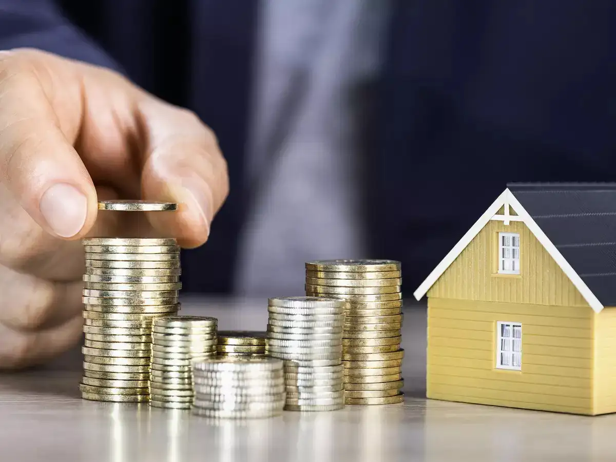 Hand placing gold coin atop stacks of coins next to a yellow toy house model on a surface