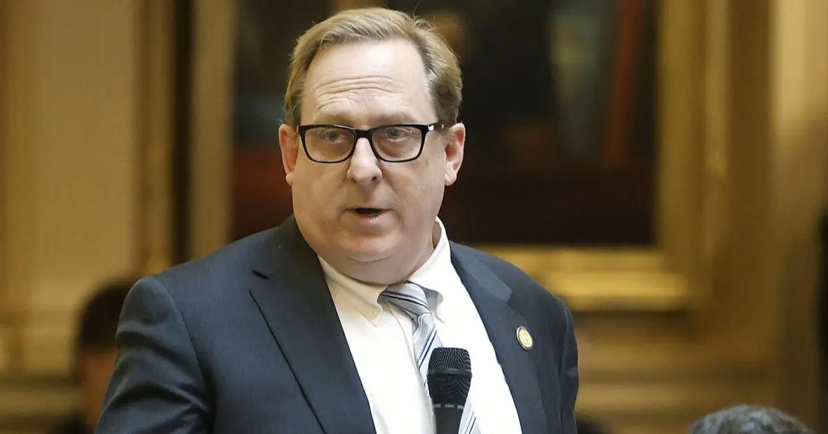 Man wearing glasses and dark suit speaking at microphone in formal interior setting
