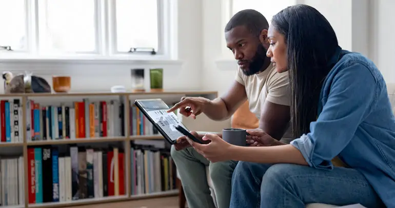 Two people sitting on a couch review documents on a tablet together in a bright living room with bookshelves