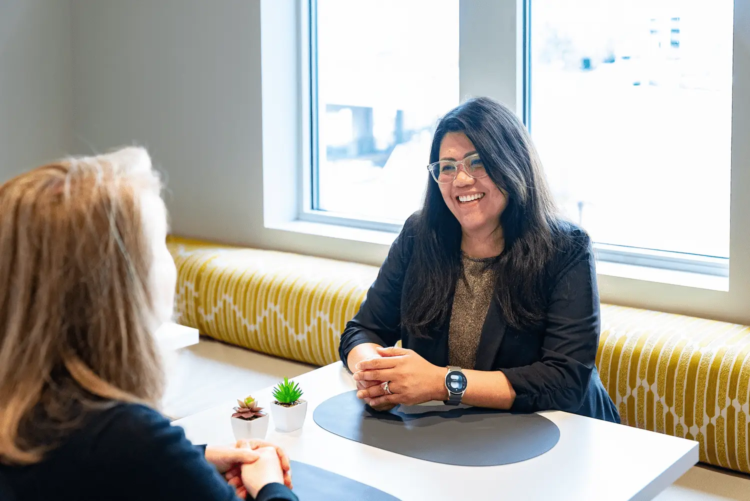 Two women sit at a table in a high-rise office, smiling during a professional conversation near large windows