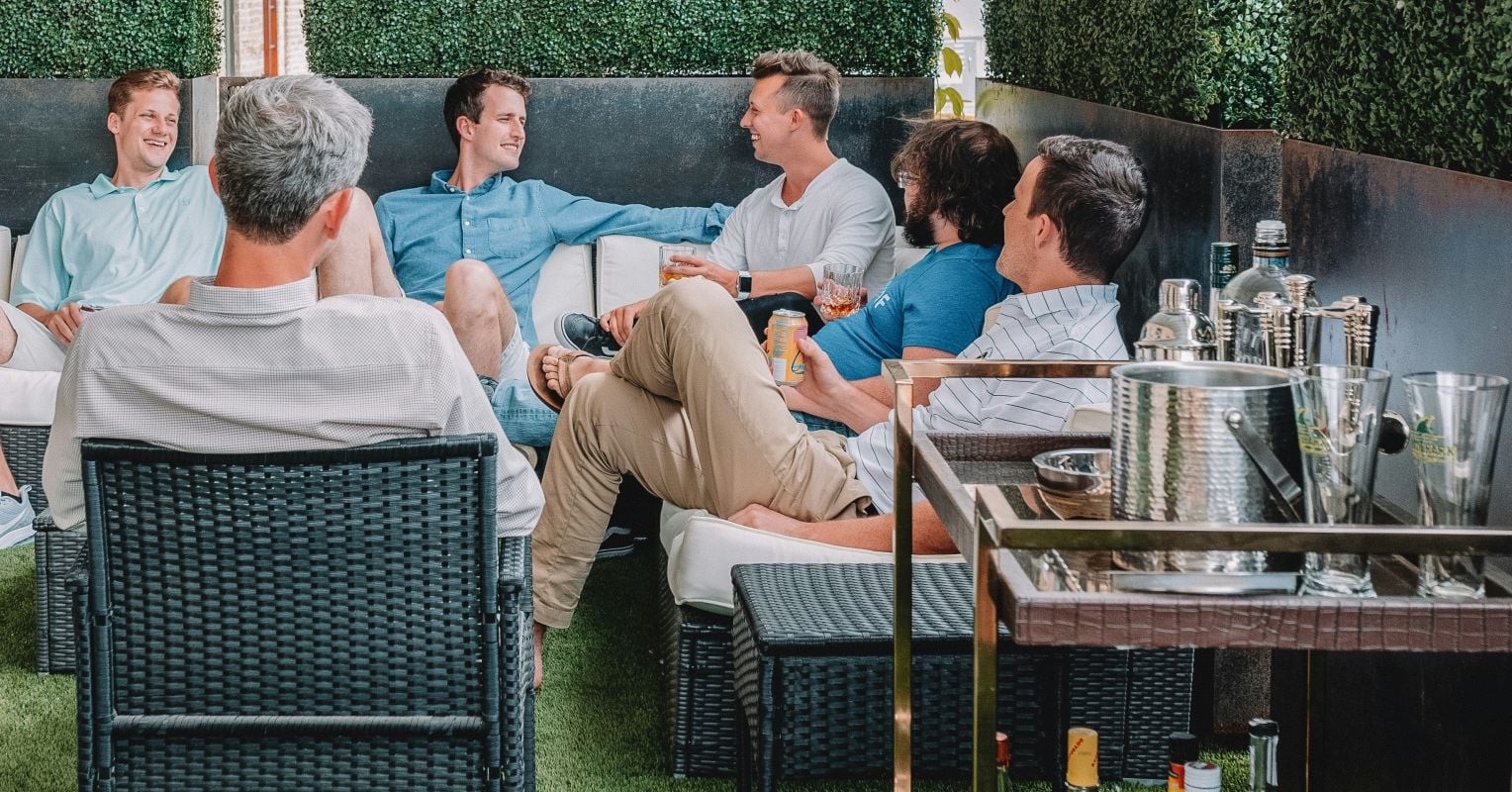 Six men sitting on outdoor furniture having drinks and conversation in a garden setting with ivy-covered walls