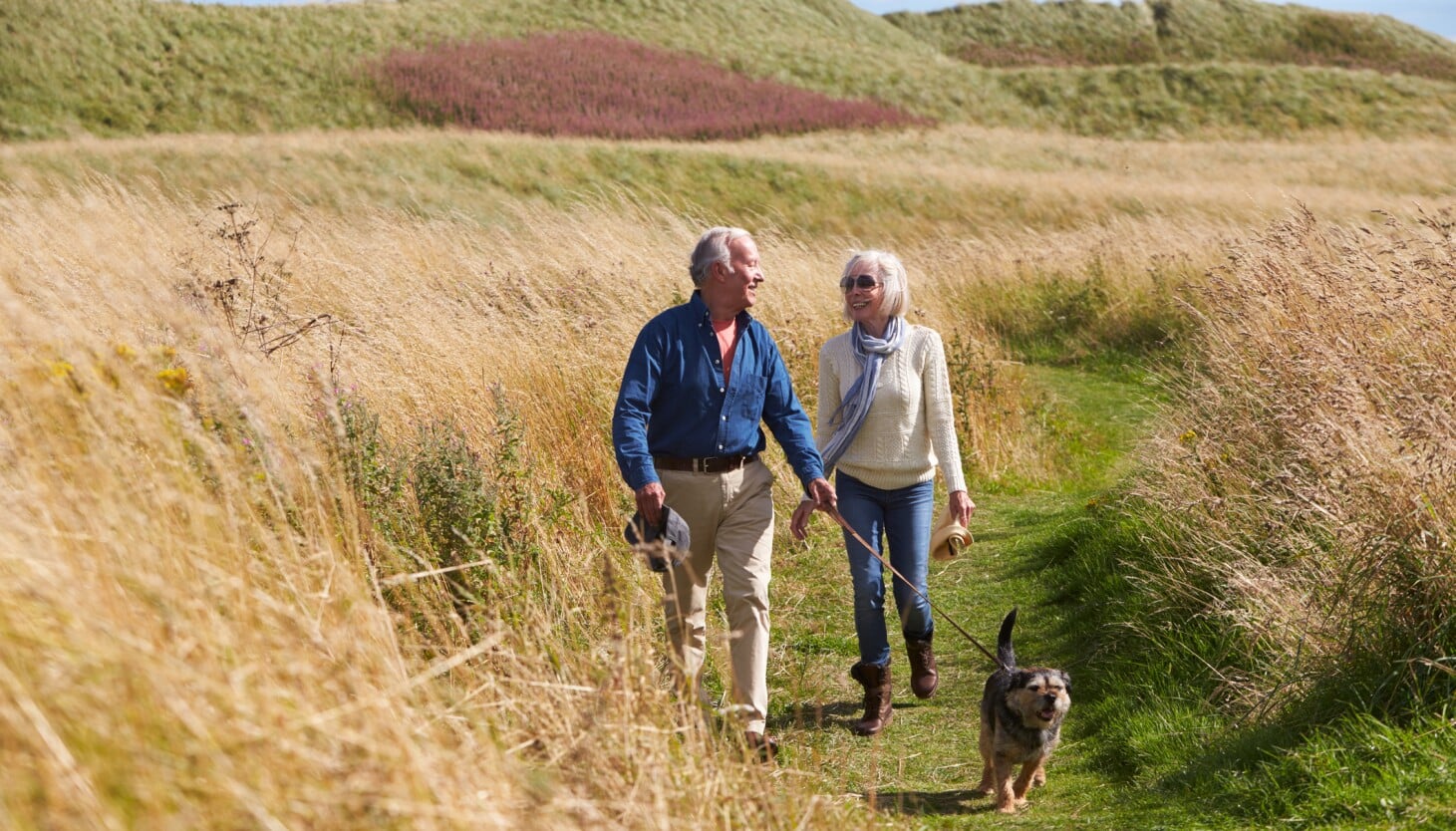 Two older adults walk on a grassy coastal path with a small dog beside them