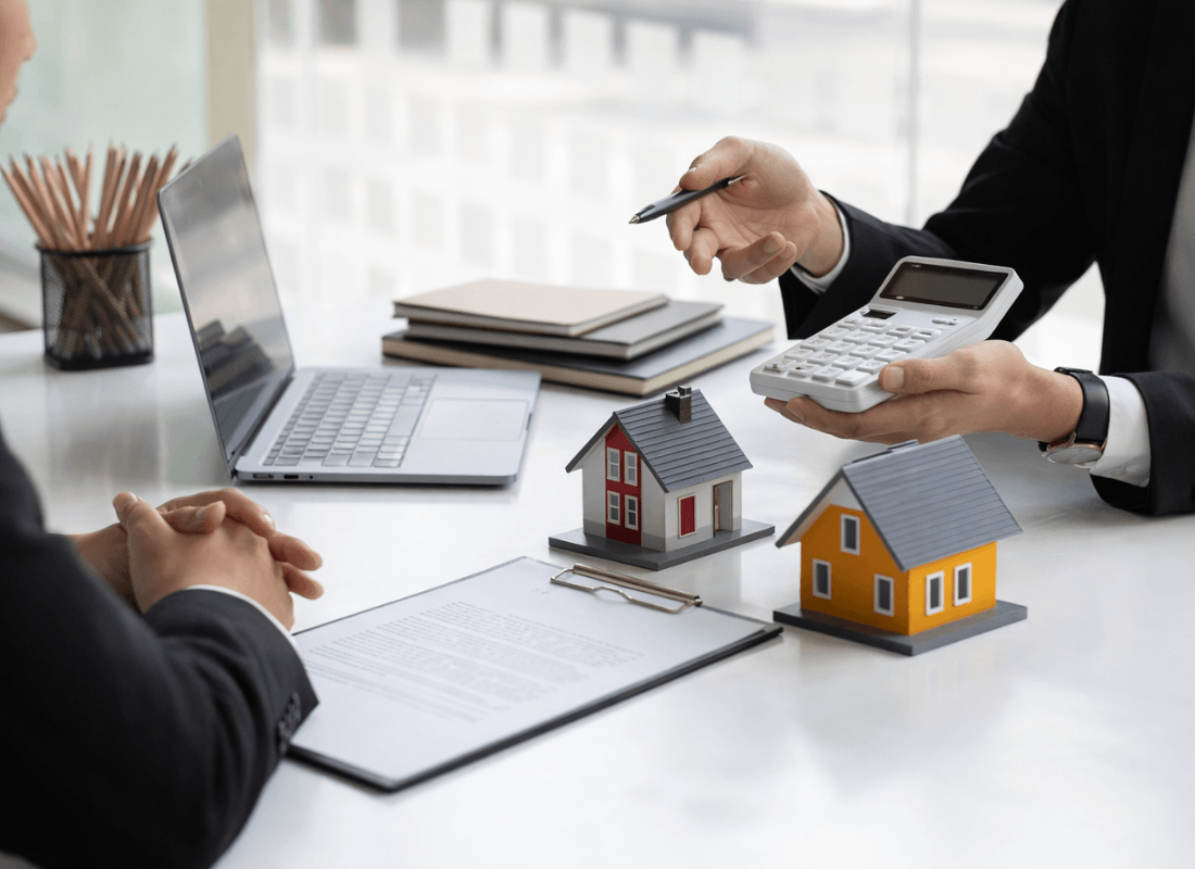 Two professionals in business attire discuss property investments at a desk with model houses and a calculator