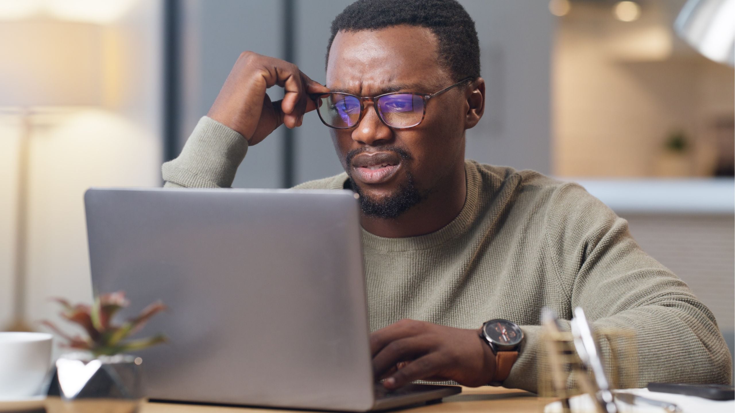 Man wearing glasses and beige sweater working on laptop at desk with plant and coffee cup visible