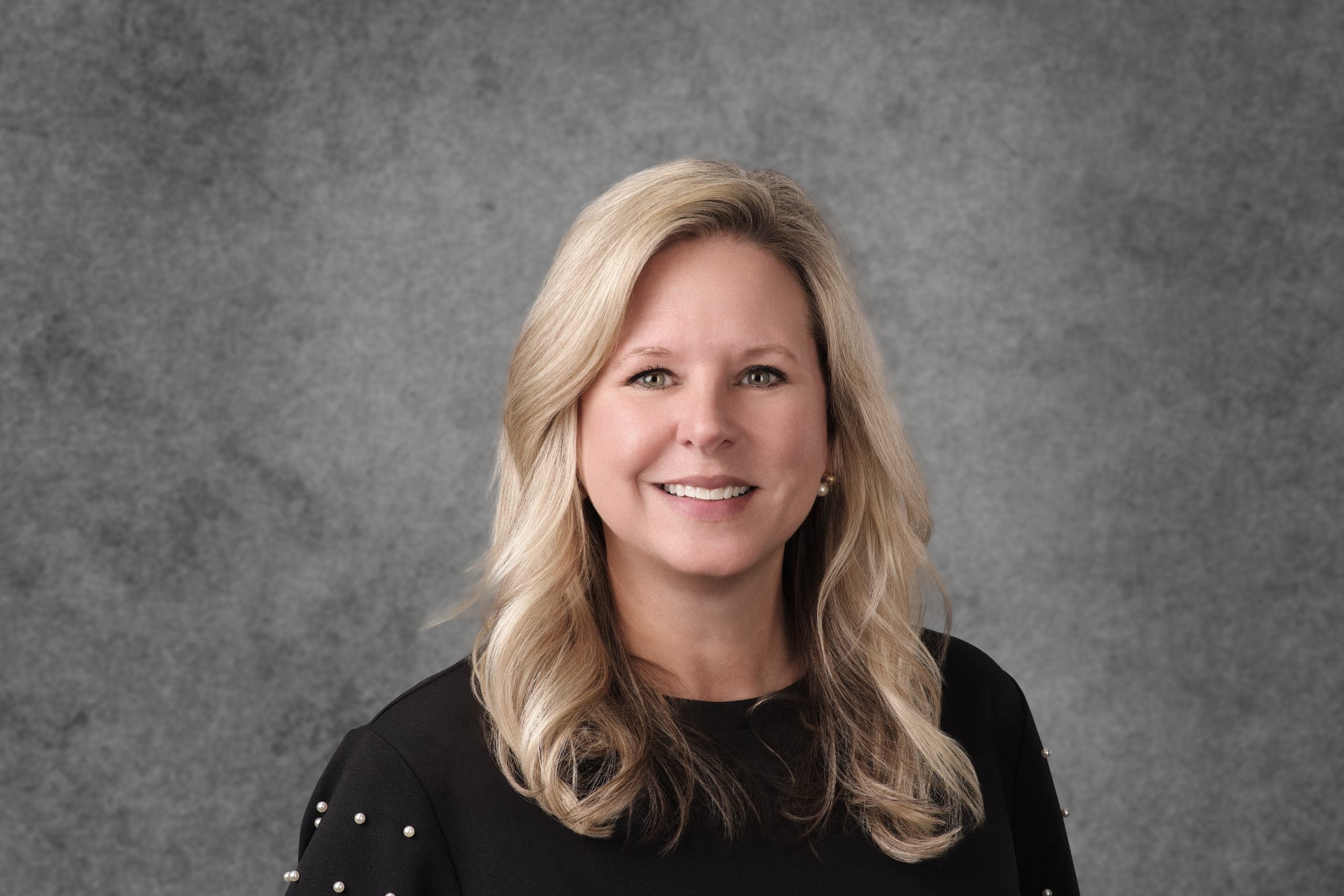 Professional headshot of a woman with shoulder-length blonde hair wearing a black top with pearl embellishments against a gray background