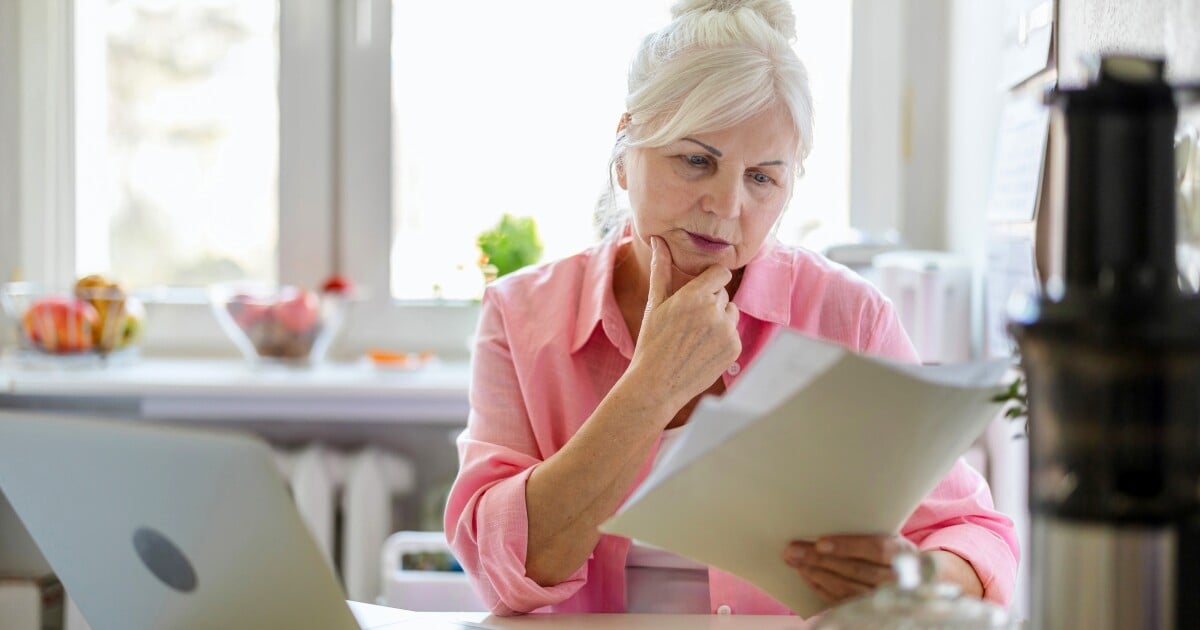 Senior woman in pink shirt reviewing document at kitchen table with laptop nearby