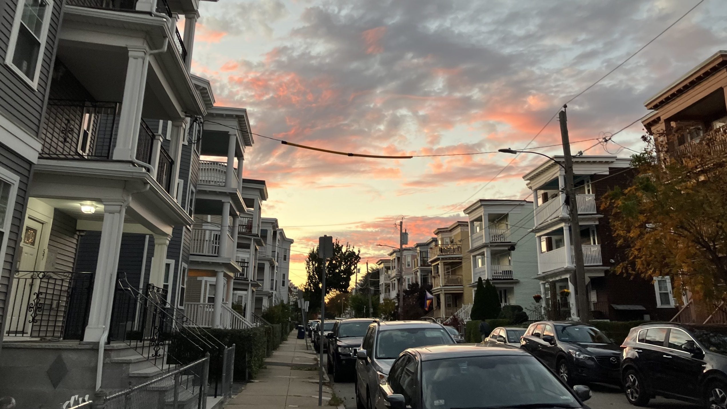 Residential street lined with multi-story buildings at sunset with parked cars and utility wires