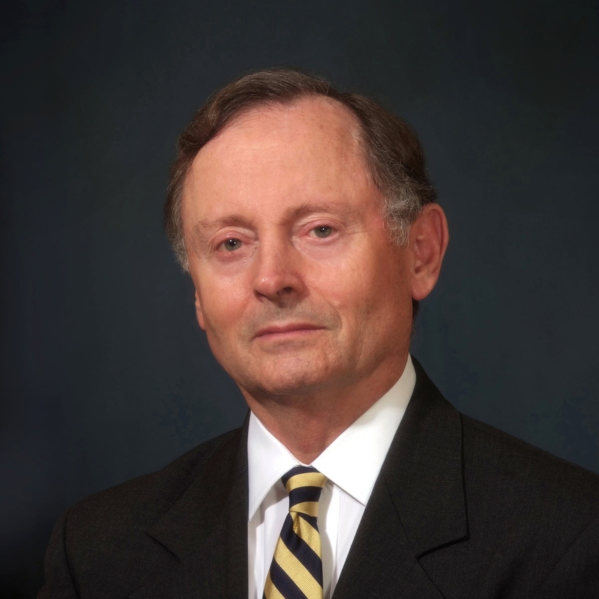 Professional headshot of a middle-aged man wearing a dark suit, white shirt, and gold and navy striped tie against a dark background
