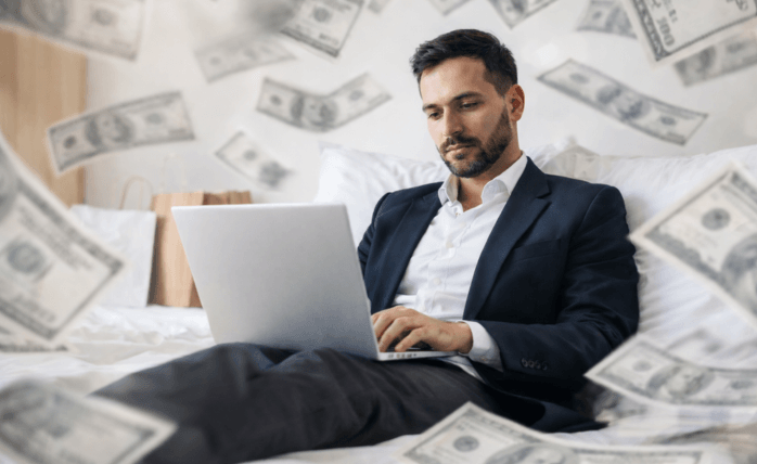 Businessman in dark blazer sitting on bed surrounded by scattered dollar bills, working on laptop