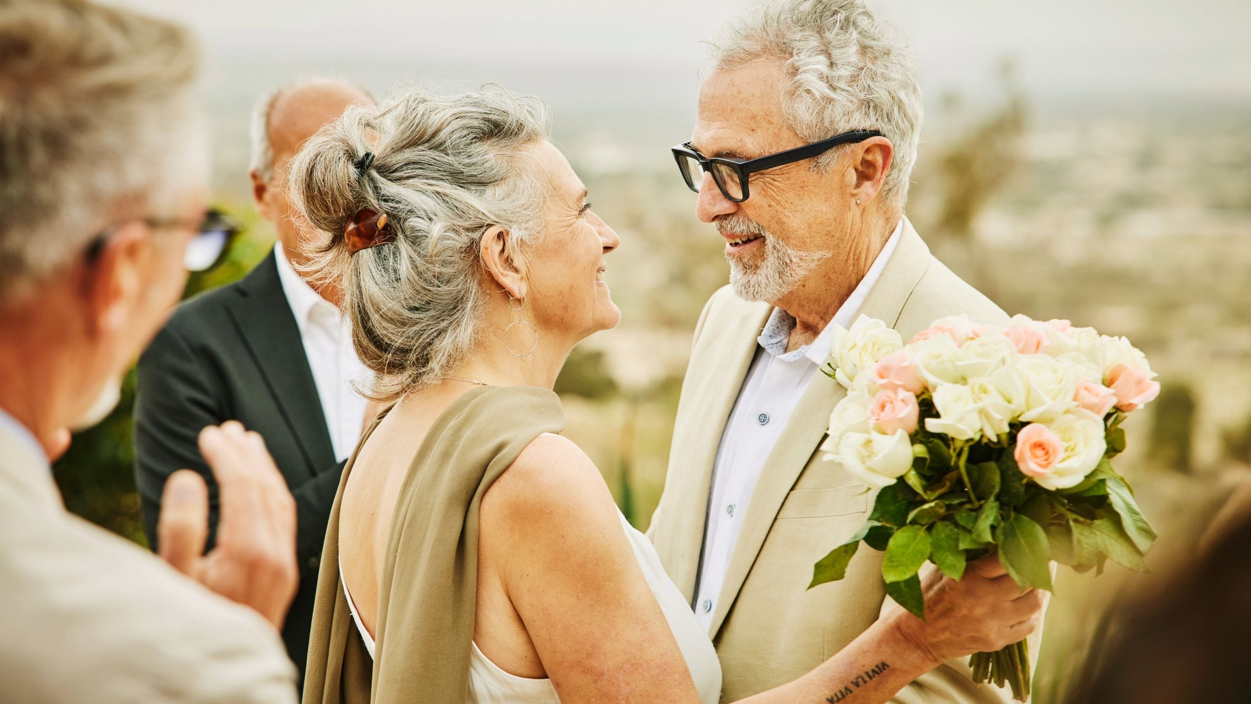 Older man and woman in beige clothing holding flowers and smiling at each other outdoors with guests nearby