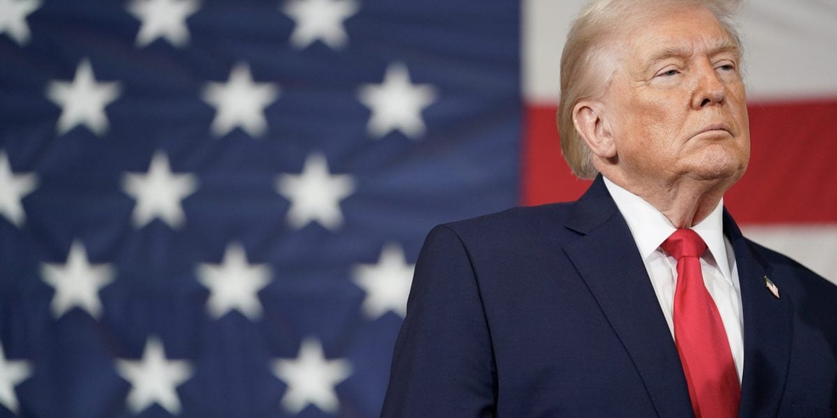 Man in dark suit and red tie standing in front of American flag backdrop
