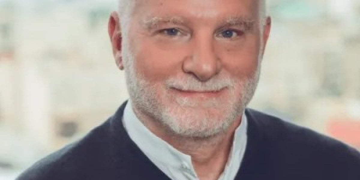Professional headshot of an older man with gray hair and beard wearing a dark blazer and light blue shirt