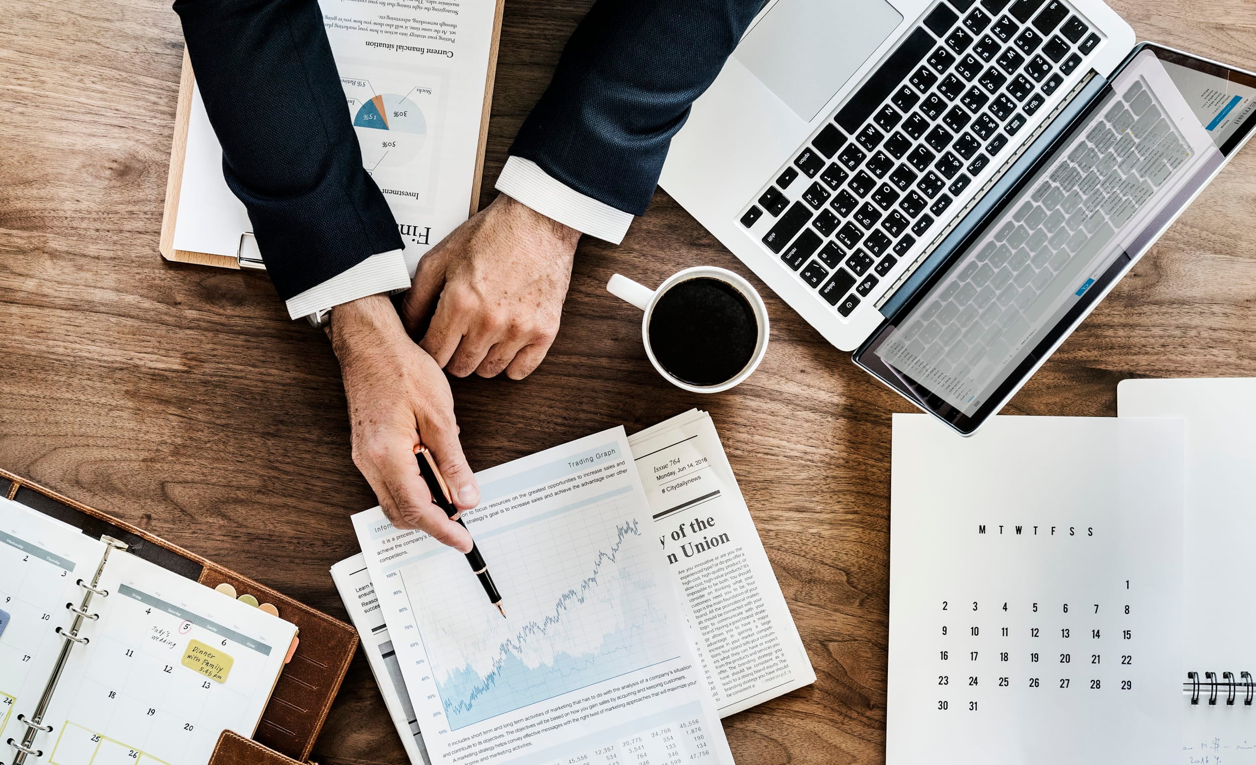Overhead view of businessman's desk with laptop, financial documents, coffee cup, calendar, and papers with charts and graphs